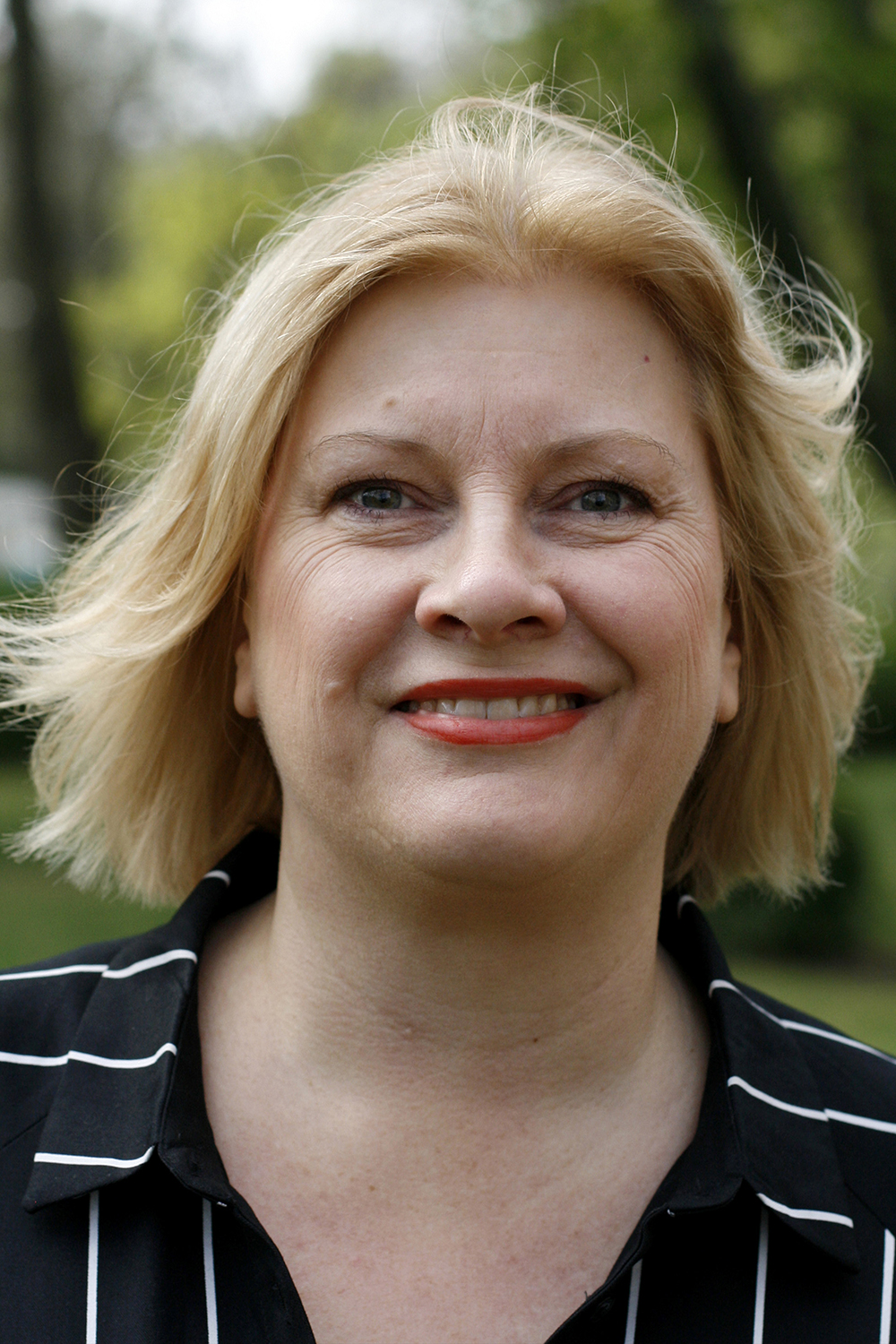 Annika: middle-aged, light-skinned woman with blonde hair. She wears red lipstick and a black shirt with bright white stripes. She is standing in front of an urban greenery and smiling.