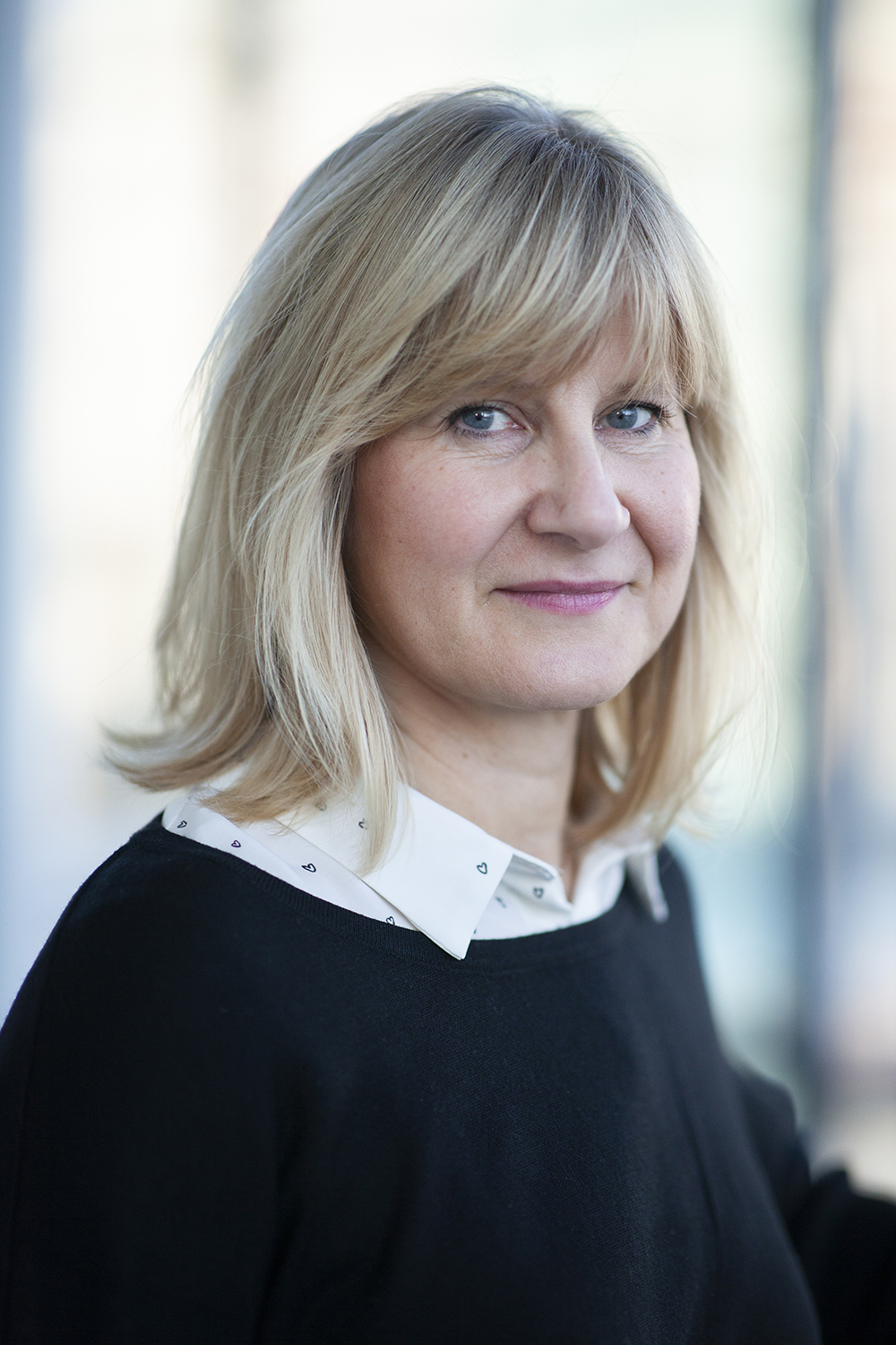 Asa: a middle-aged, light-skinned woman with blonde short hair and blue eyes. She smiles at the camera and wears a white shirt with small heart shaped details in black. On top of the shirt, she wears a think black pull over. She stands in a well lighten room that is blurred by the camera lens into blue, white and yellow colors.