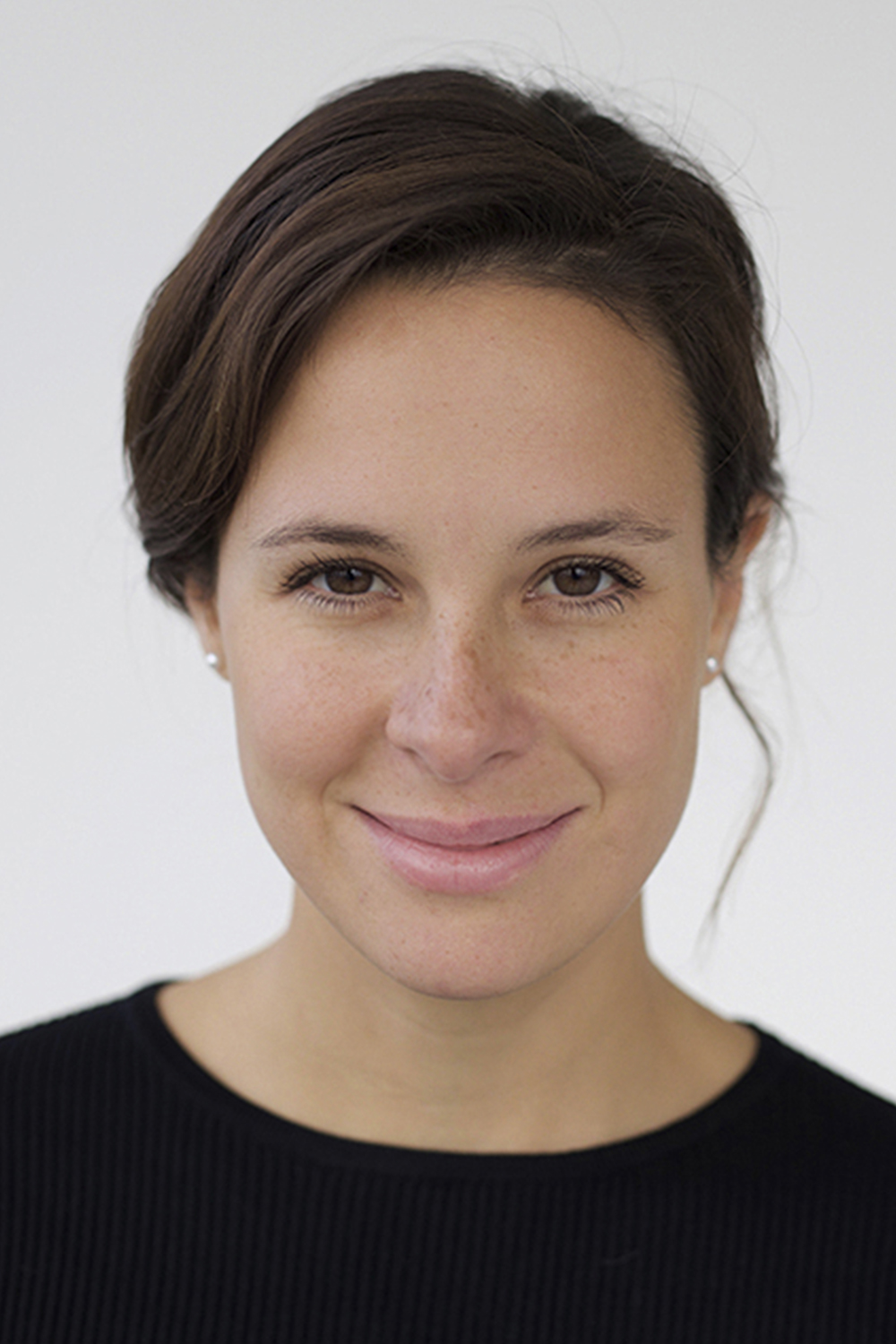 Barbara: a 30-year-old olive-skinned woman with brown eyes and hair. She has freckles on her nose and cheeks. She wears a simple black long sleeve t-shirt and small pearl earrings. Her hair is wavy and tied at the back. She is at her friend's photography studio who took the photo (April Vedby Kristiansen).