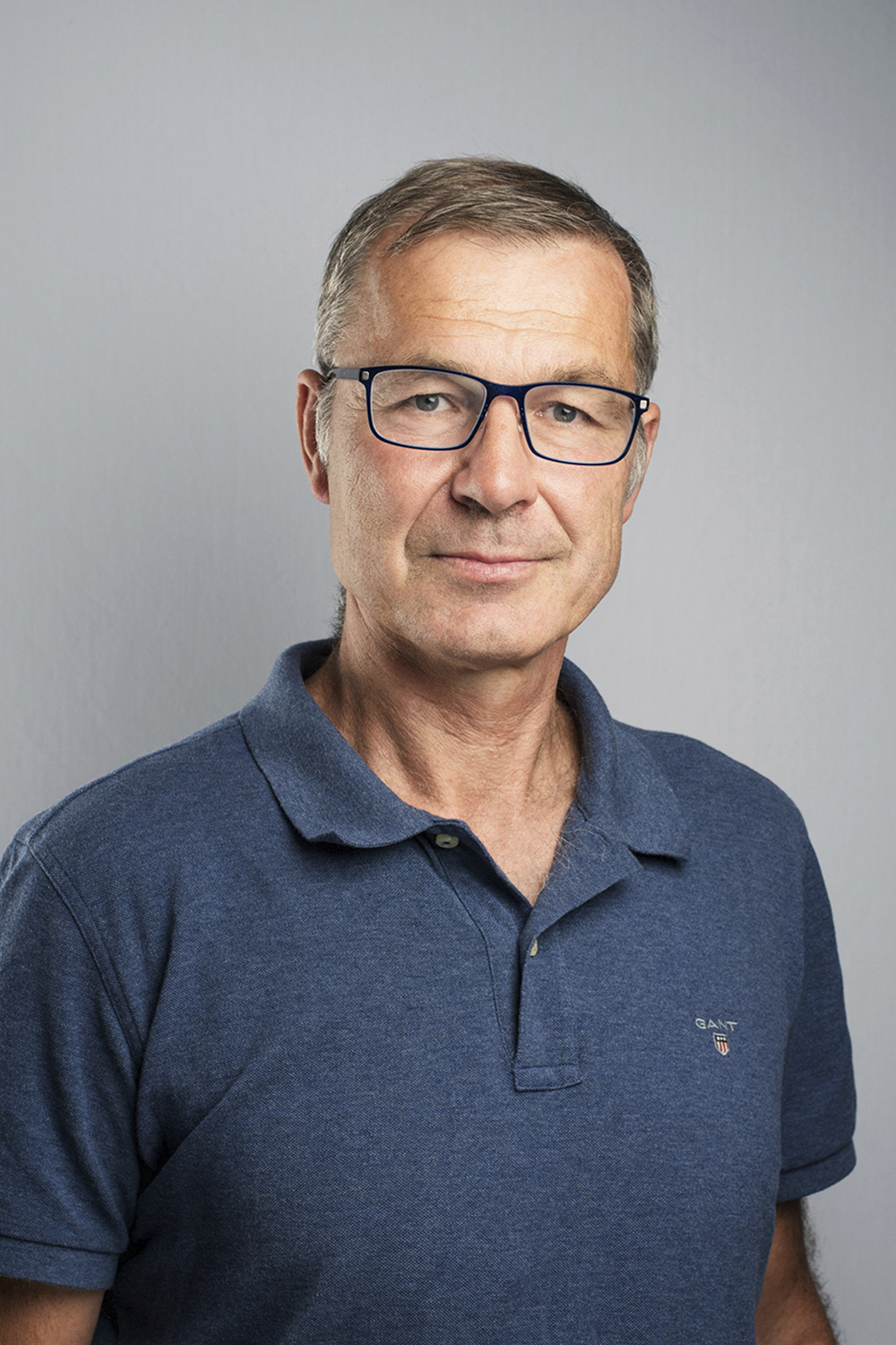 Carl: a middle-aged, light-skinned man with blue eyes and short grey hair wearing squared glasses. He wears a Gant blue polo and stands in a studio with a dark grey background.