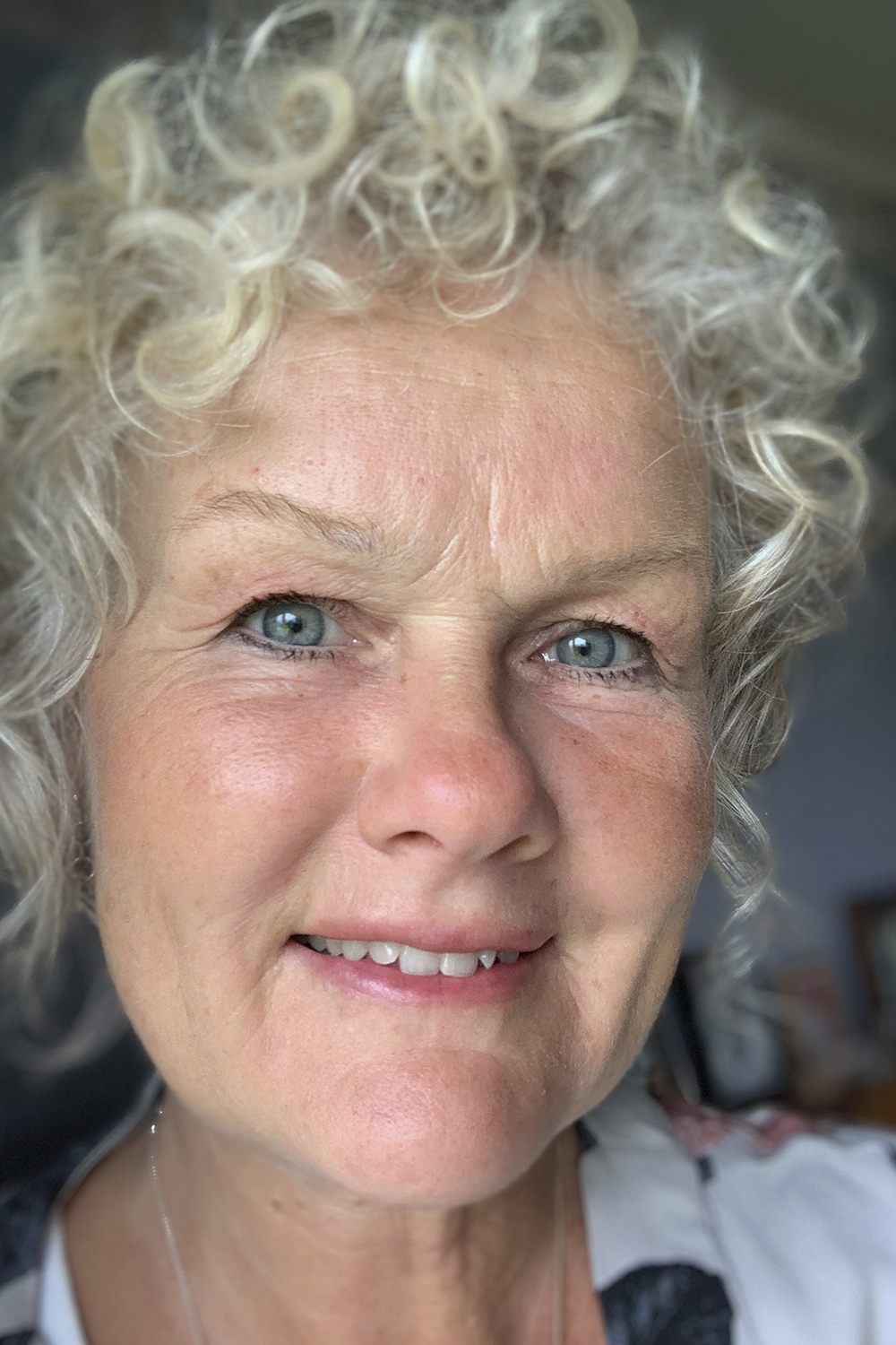 Catarina: a middle-aged, light-skinned woman with blue eyes and curly white hair. She is standing very close to the camera inside a room that is hard to see due to the image framing. She wears a silver necklace and a white shirt with flower patters.