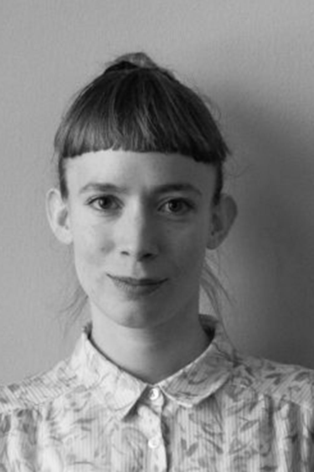 Irina: a black and white photograph of Irina, a young light-skinned woman with brown hair, she is looking at the camera smiling against a white wall. She has a sharp fringe, and her hair is tied at the back. She is wearing a white shirt with flower patterns