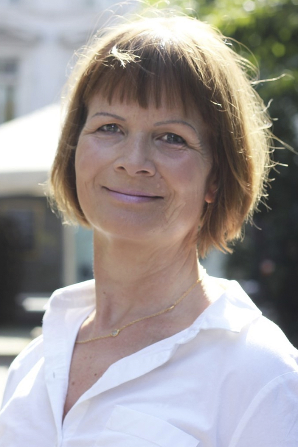 Michala: a middle-aged, light-skinned woman with light brown hair and a fringe. She wears light pink lipstick, a white shirt and a golden necklace. Behind her, we see a blurred image of a sunny facade surrounded by trees.