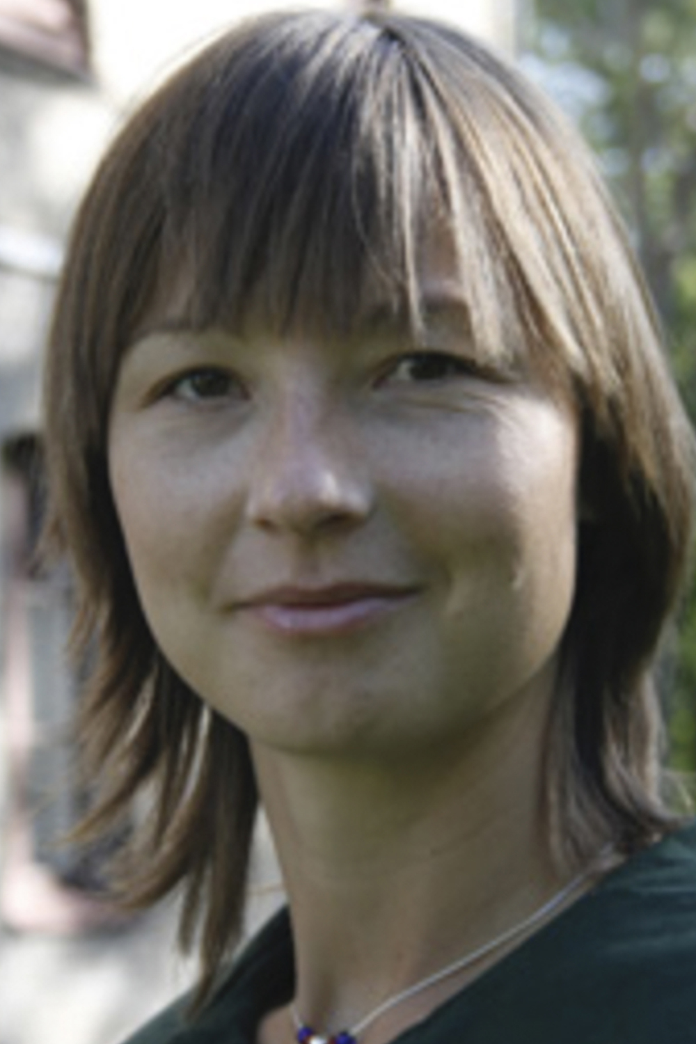 Mona: a young light-skinned woman with brown hair and eyes. She wears a necklace of white and blue colours and a dark green shirt. She is standing somewhere outside, presumably in Gothenburg.