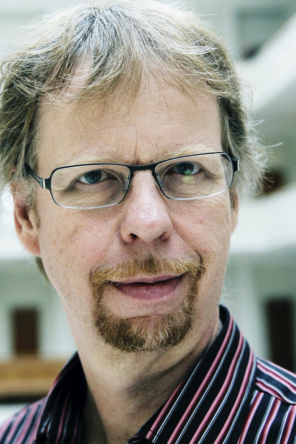 Søren: a middle-aged, light-skinned man smiling at the camera with rectangular glasses, a beard and blonde-greyish hair. He seems to be standing inside a well-lighted room that may be a public building such as a university or a citizen service center.