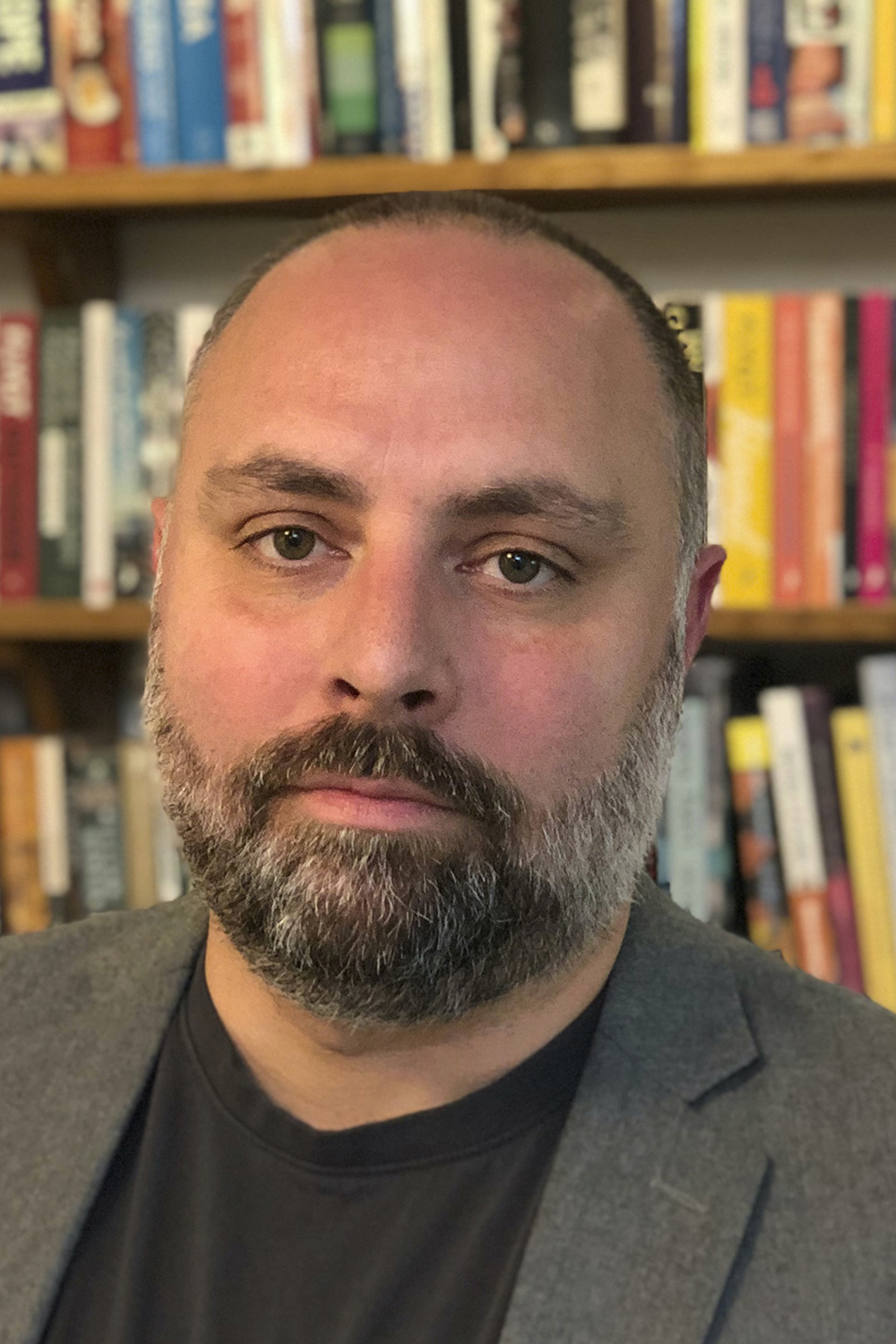 Thomas: a man in his 40s with light-skin, brown eyes and white hair and beard. He is looking at the camera with a calm posture. Behind him an overflowing library of colourful books. He wears a black t-shirt and a grey blazer.